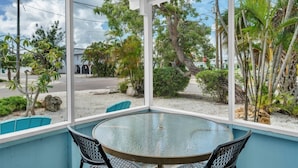 A glass-enclosed patio with a round table and two chairs, overlooking a street with palm trees and a stop sign. - Minnow 1 1 Bedroom Cottage by RedAwning (Anna Maria)