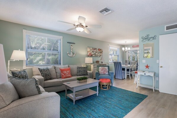 A living room with a sofa, coffee table, and a dining area with a chair and table. - Beach Breeze Hideaway 2 Bedroom Villa by RedAwning (Holmes Beach)