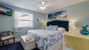 A bedroom with a bed, a chair, a desk, a ceiling fan, and artwork on the walls. - Beach Breeze Hideaway 2 Bedroom Villa by RedAwning (Holmes Beach)