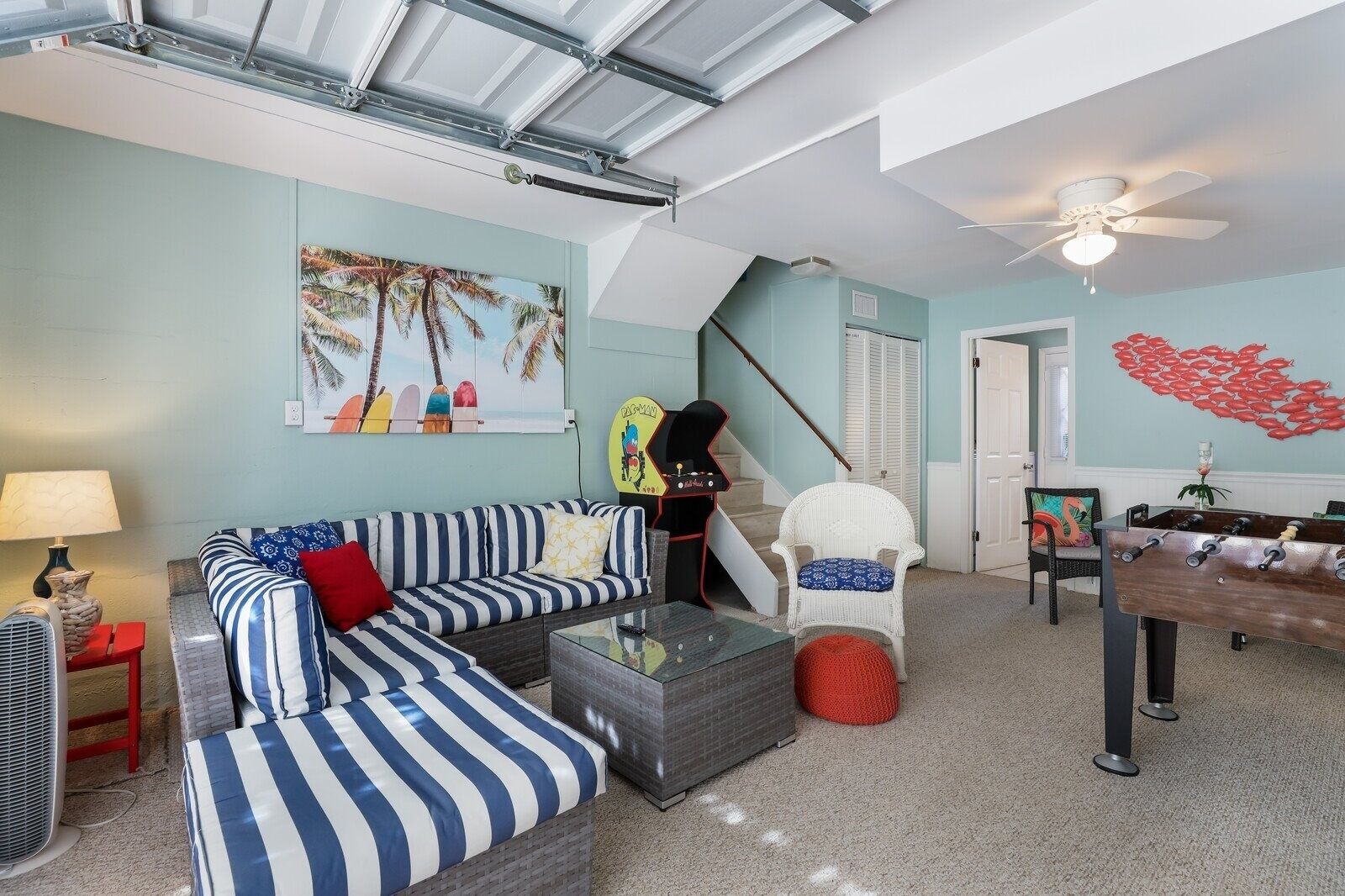 A living room with a sofa, a chair, a foosball table, and a painting of palm trees.