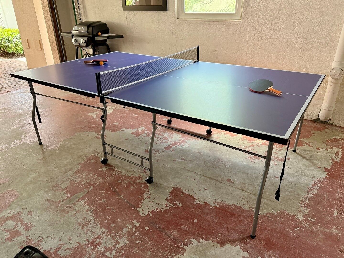 Una mesa de ping pong con red en una habitación de piso de concreto y una parrilla al fondo.