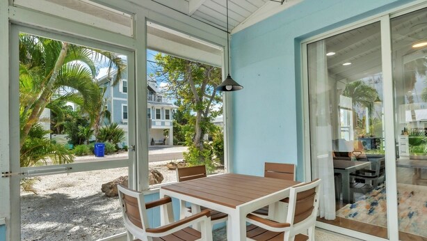 A covered outdoor dining area with a table and chairs, large windows, and a view of a residential neighborhood. - Minnow 2 2 Bedroom Villa by RedAwning (Anna Maria)
