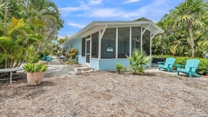 A small, light blue building with a covered porch, surrounded by tropical plants and outdoor seating. - Minnow 2 2 Bedroom Villa by RedAwning (Anna Maria)