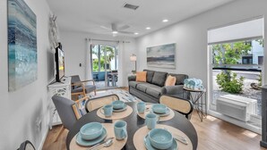 A modern living room with a gray sofa, a dining table set with blue plates and cups, and a large window with a view of the outdoors. - Minnow 2 2 Bedroom Villa by RedAwning (Anna Maria)