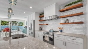 Una cocina moderna con armarios blancos, una isla central, electrodomésticos de acero inoxidable y estanterías de madera.