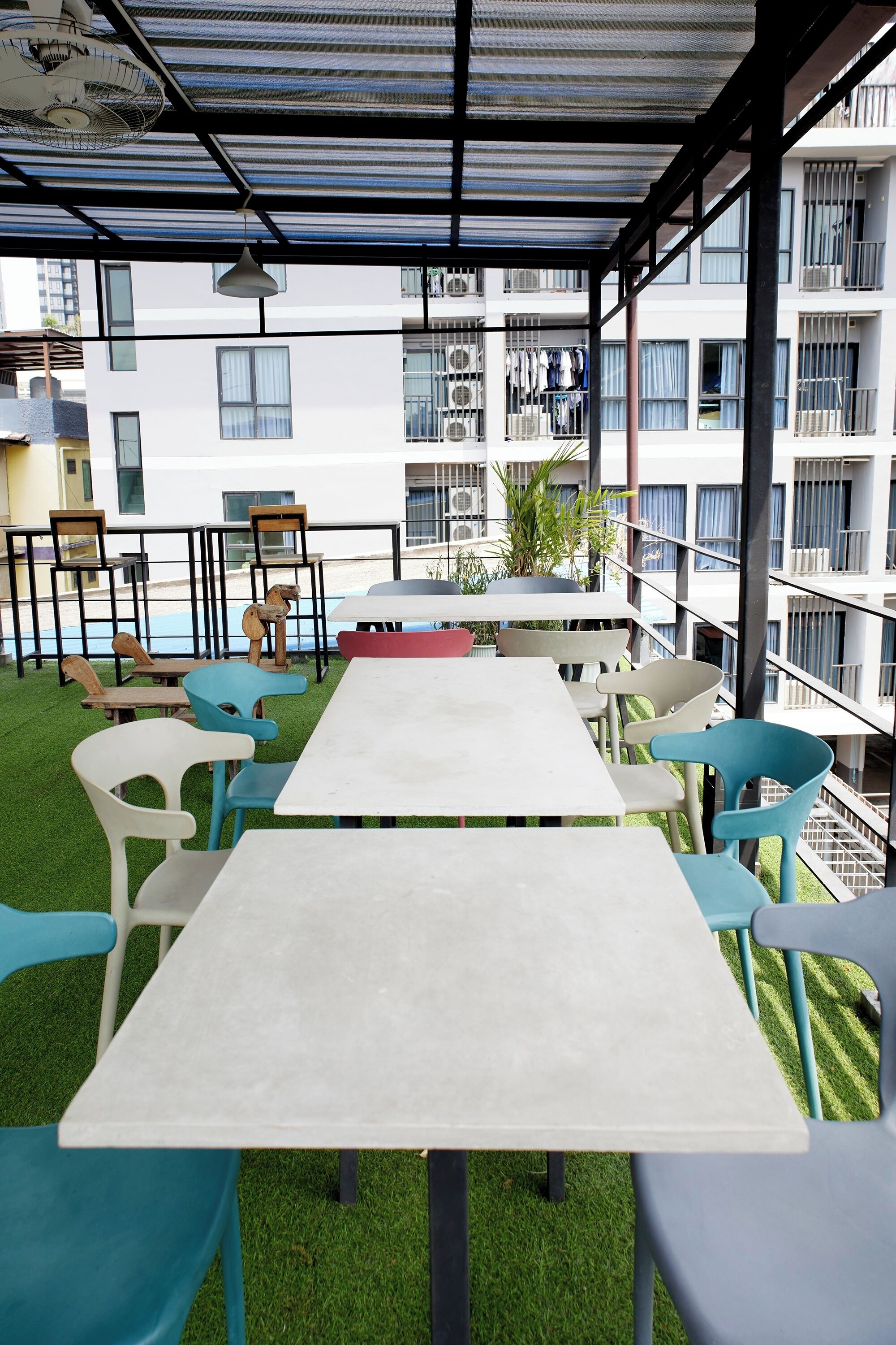 A covered outdoor seating area with a long table and colorful chairs, overlooking a residential building with balconies and air conditioning units.