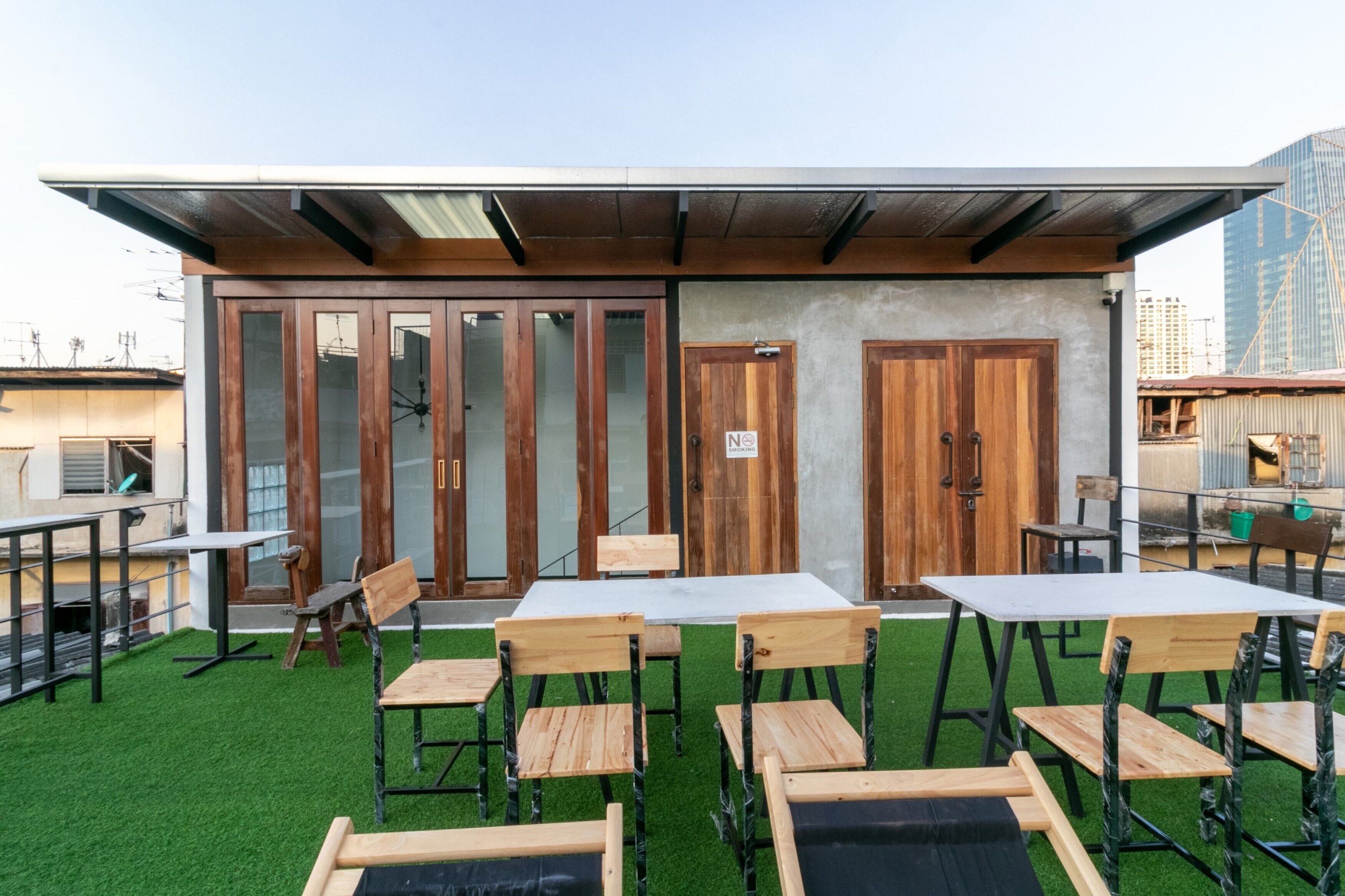 Un espacio al aire libre con mesas y sillas de madera, un muro de hormigón y una estructura de techo metálico.