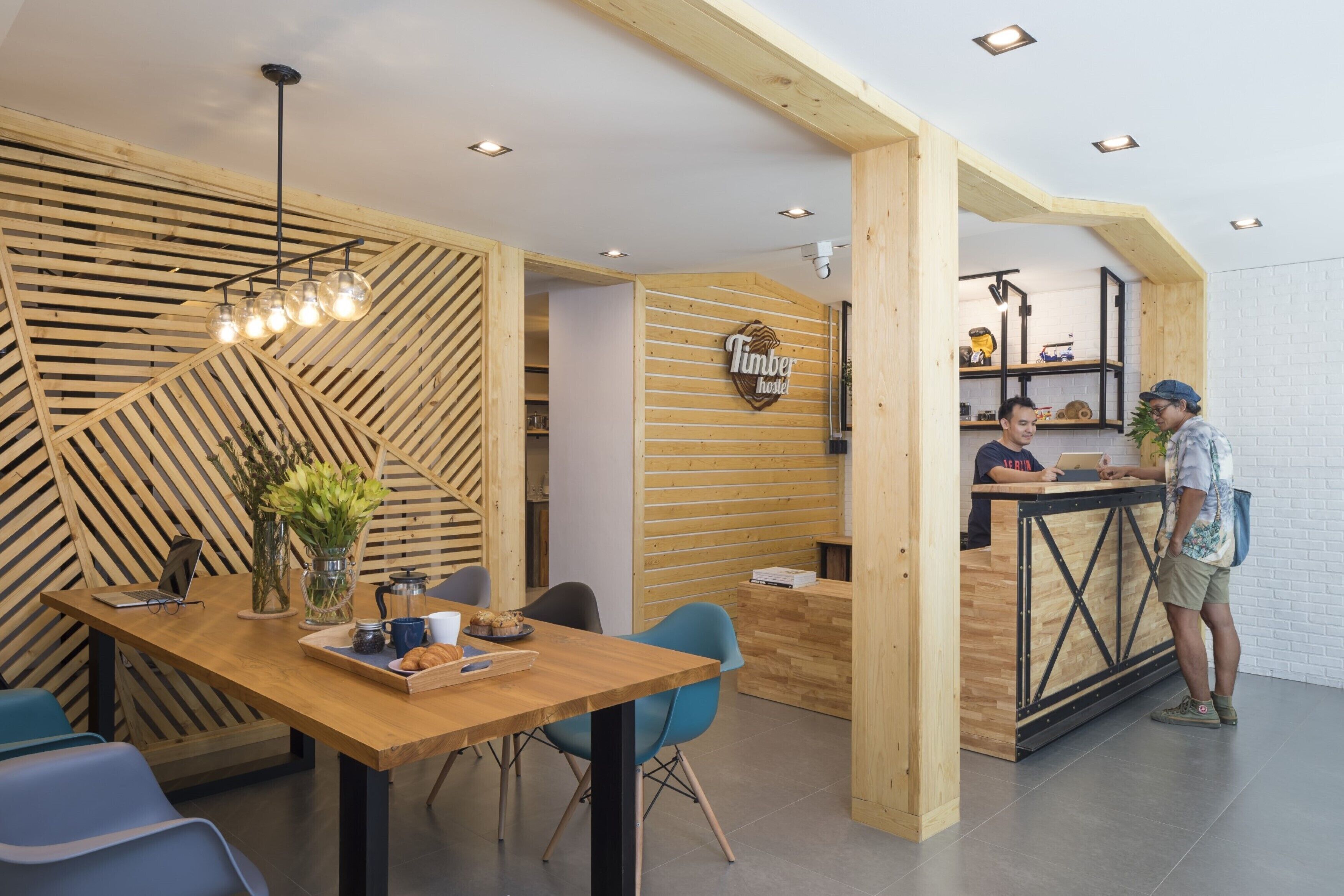 A modern interior with a wooden reception desk, a seating area with a laptop and a tray of pastries, and a visible sign for 'Timber Hostel'.