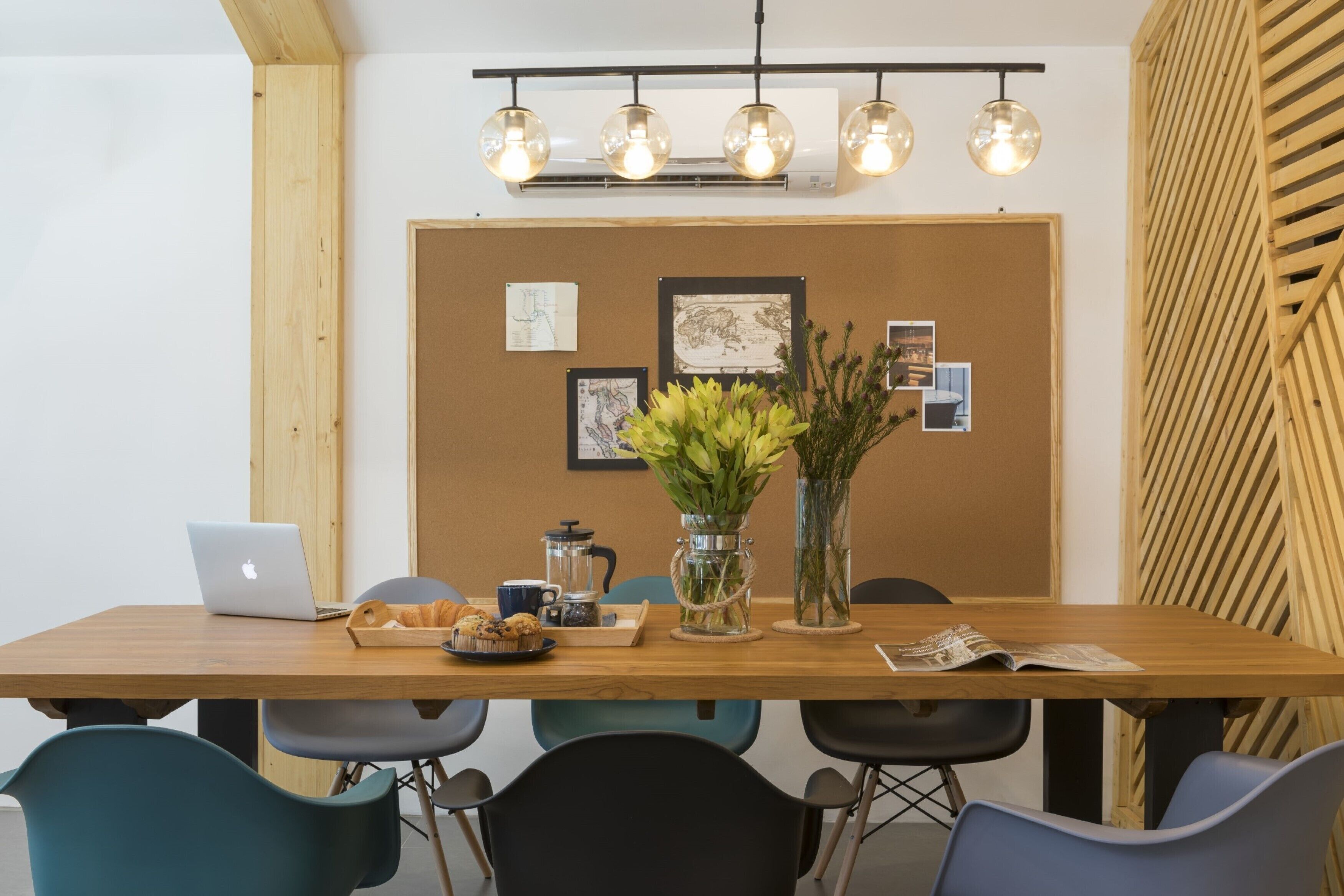 A conference room with a wooden table, chairs, a laptop, a coffee pot, and a bulletin board with pictures and a vase of flowers.