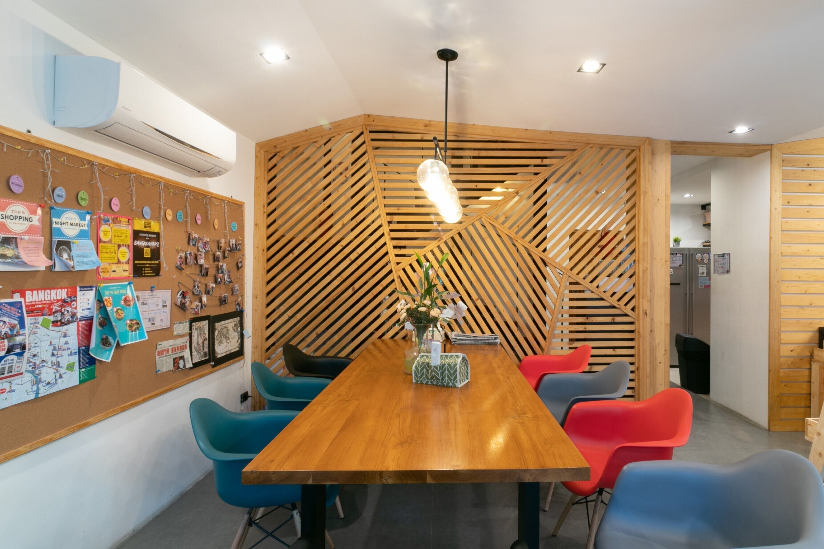 A modern meeting room with a wooden table, colorful chairs, a bulletin board with various notices, and a slatted wooden partition.