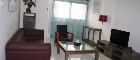 A living room with a sofa, a glass coffee table, a TV stand, a lamp, and a potted plant.