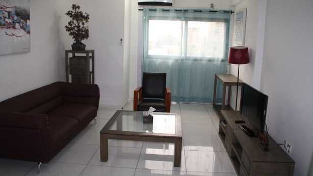 A living room with a brown sofa, a glass coffee table, a wooden cabinet, a TV, and a red lamp. - L'Almamya Hôtel & Résidence (Conakry)