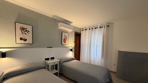 A hotel room with two beds, a bedside table, a wall-mounted air conditioner, and a window with curtains. - Hostal Bahía (Cádiz)
