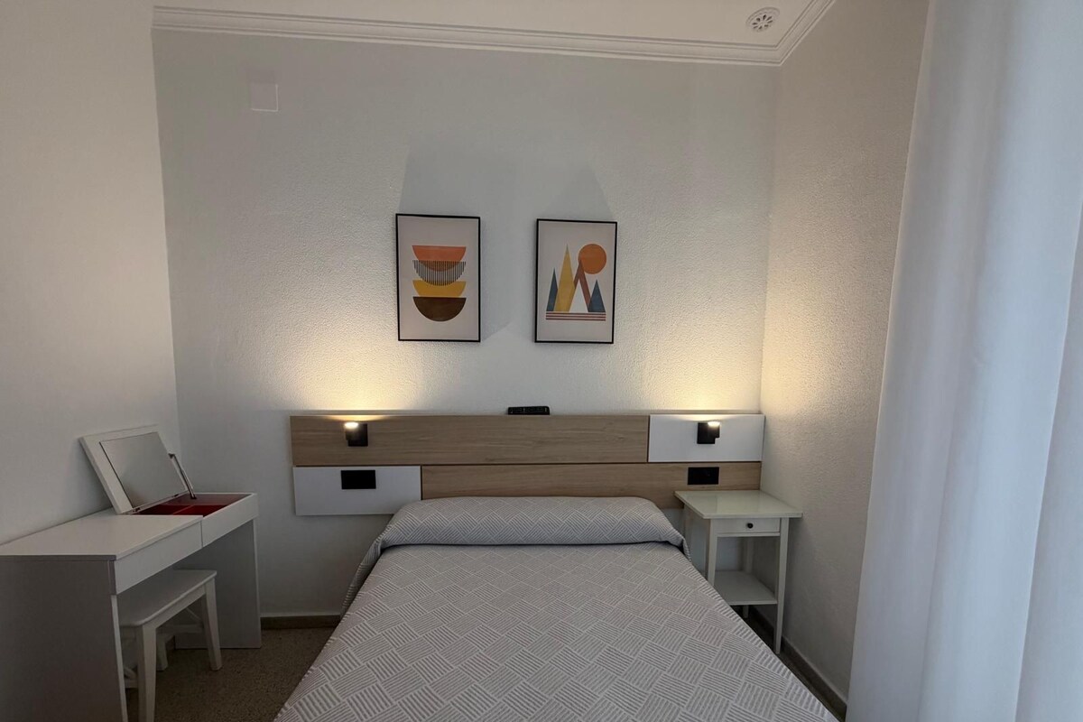 A bedroom with a bed, bedside tables, a desk, and two framed artworks on the wall.