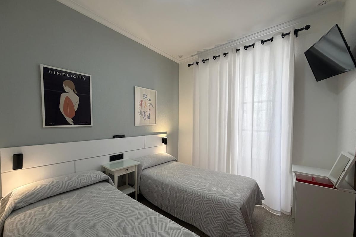 A hotel room with two beds, a flat-screen TV mounted on the wall, and a window with white curtains.