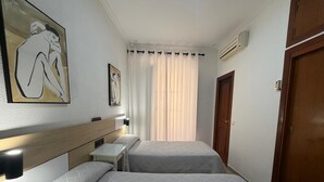 A hotel room with two beds, a wooden headboard, a bedside table, a wall-mounted air conditioner, and a large window with curtains. - Hostal Bahía (Cádiz)