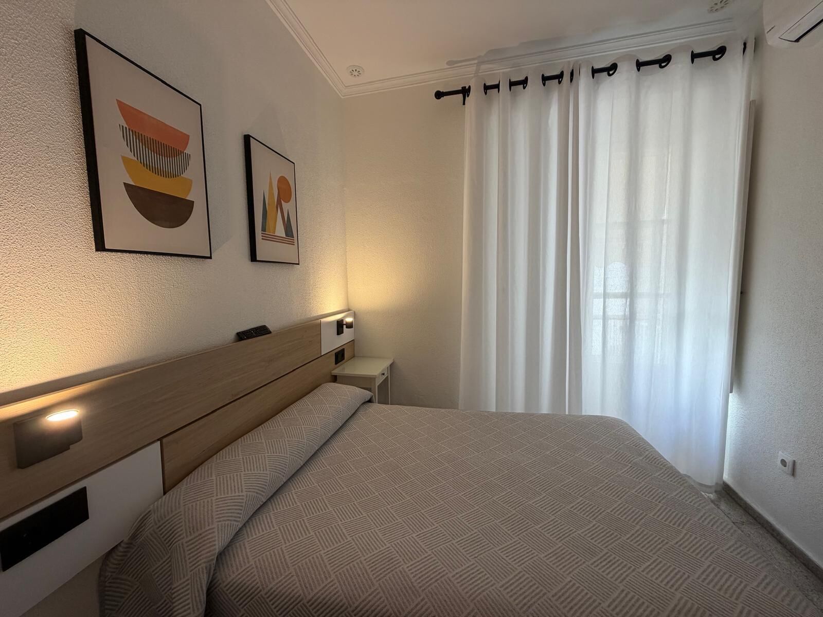 A hotel room with a bed, a nightstand, a wall-mounted lamp, and two framed artworks.