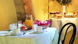 Una mesa de desayuno dispuesta con tazas, una jarrón y un bol de fruta, con una lámpara y flores al fondo.