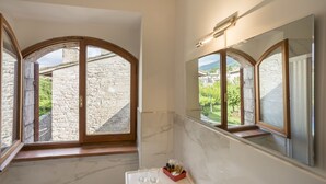 Un baño con paredes de piedra, un lavamanos de mármol y amplias ventanas de arco que dan a un edificio de piedra y vegetación.