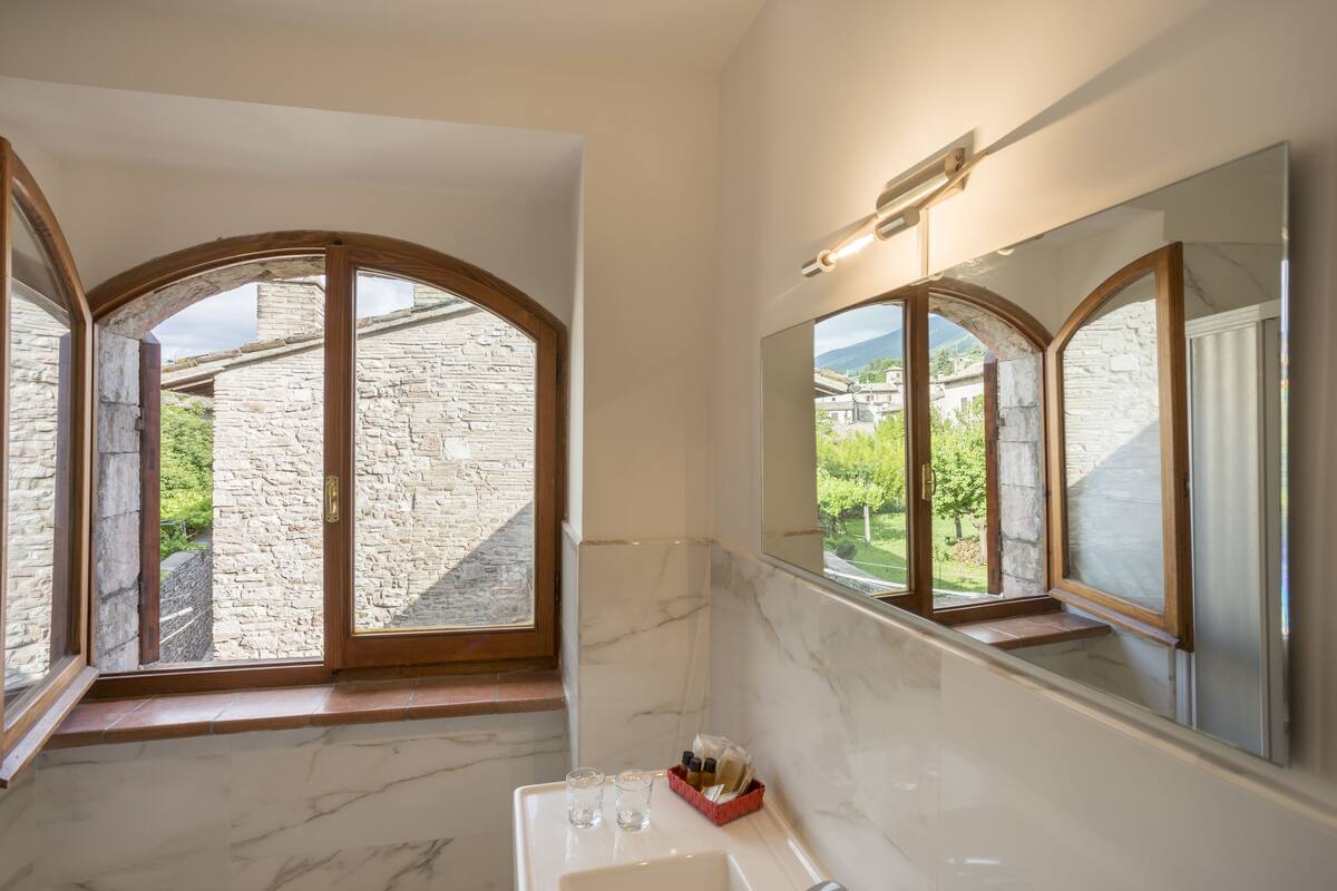 A bathroom with stone walls, a marble sink, and large arched windows overlooking a stone building and greenery.