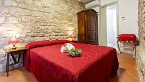 Una habitación con pared de piedra, una cama con colcha roja, un armario de madera, una mesita de noche con una lámpara y una maleta sobre un atril.
