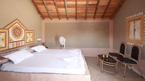 Une chambre avec un plafond en bois, un lit recouvert de draps blancs, une table de chevet avec un ventilateur et deux chaises.