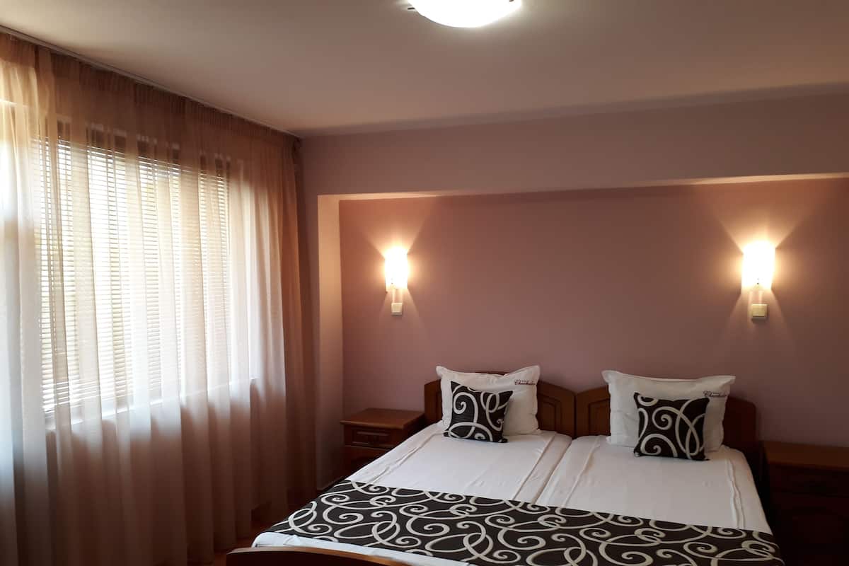 Ein ordentlich bezogenes Bett mit einem gemusterten Überwurf, zwei Nachttischlampen und einem Fenster mit durchsichtigen Vorhängen.