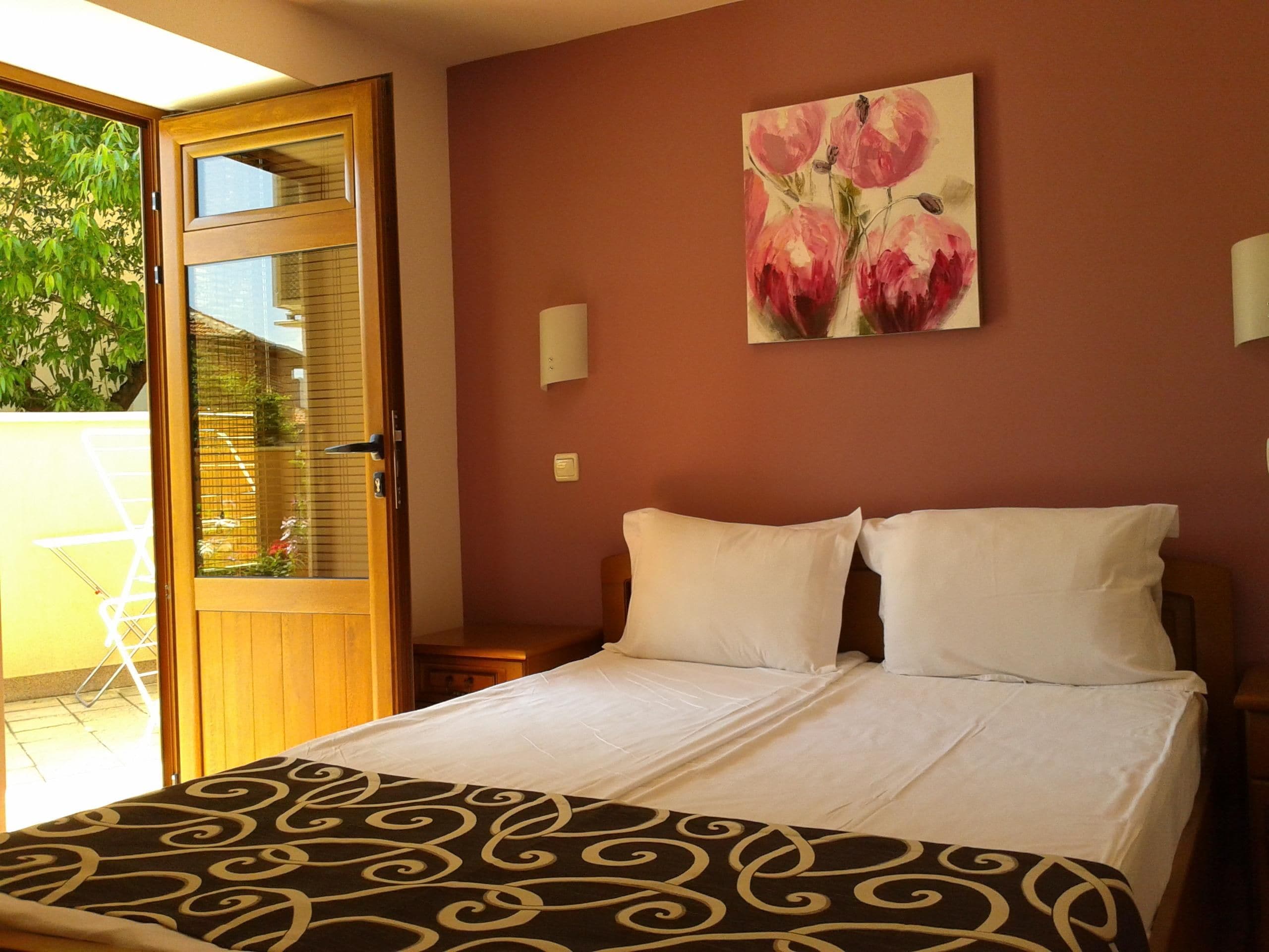 Una habitación de hotel con una cama, un cuadro de flores en la pared, una ventana con vista a follaje y una puerta.