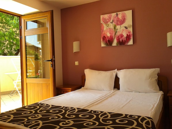 A hotel room with a bed, a painting of flowers on the wall, a window with a view of greenery, and a door. - Chuchulev (Sozopol)