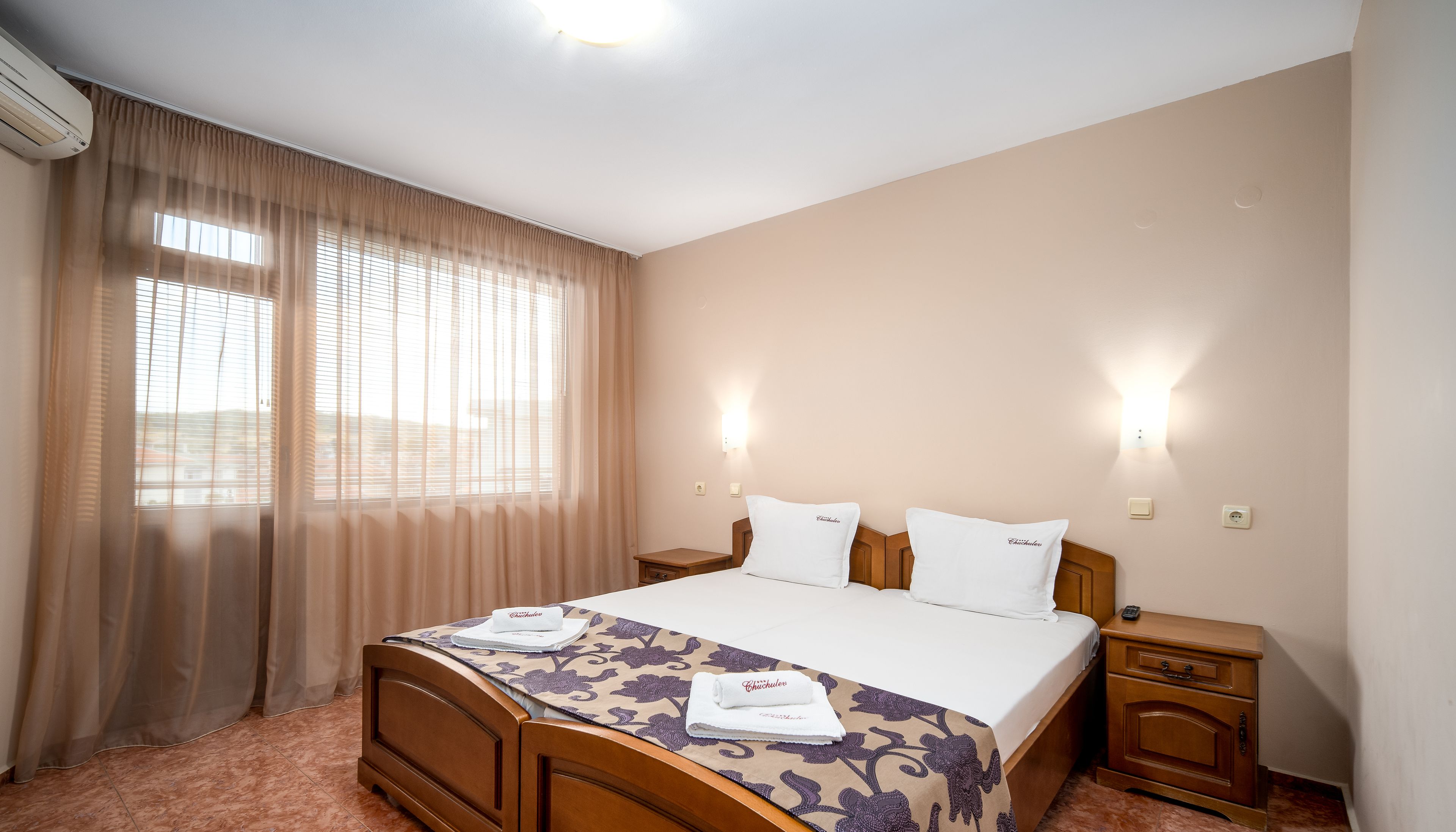 A hotel room with a bed, nightstands, a window with curtains, and a wall-mounted light fixture.