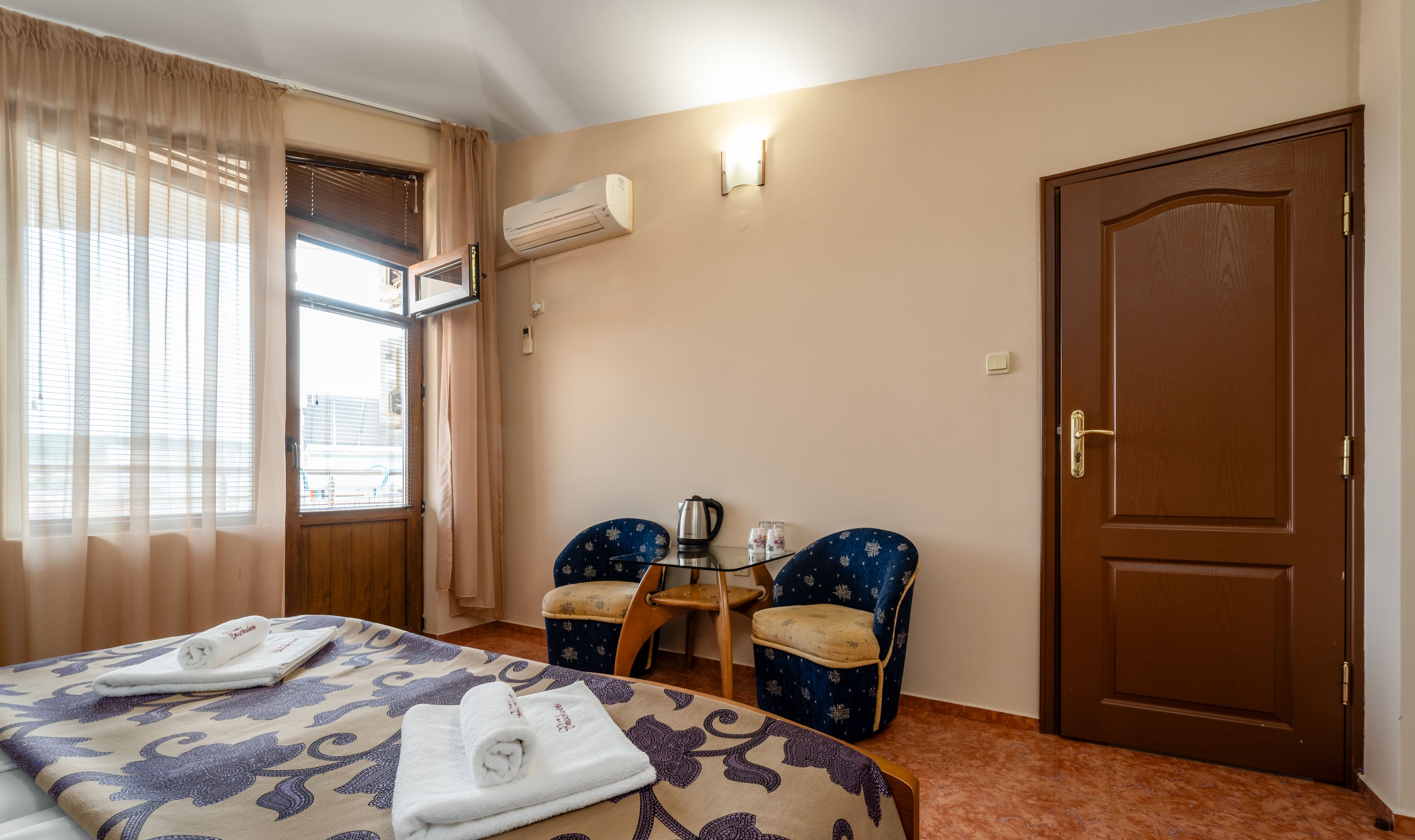 Una habitación de hotel con una cama, dos sillas, una mesita con una tetera y una puerta.