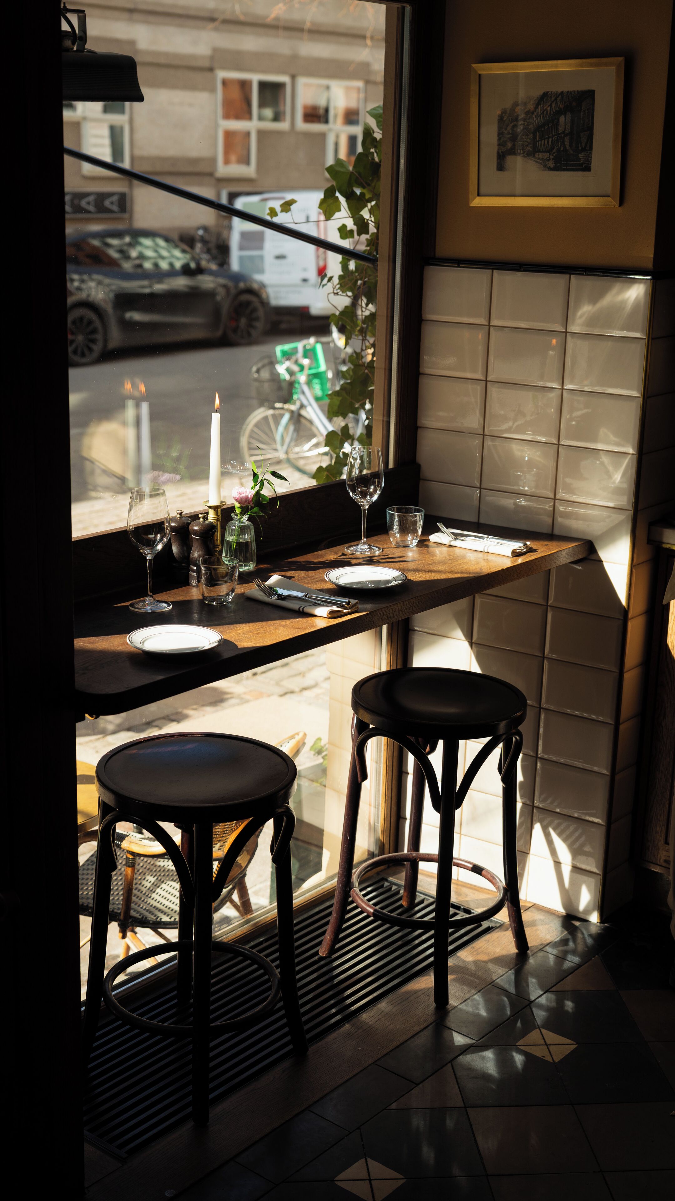 Un coin repas avec une table dressée pour deux personnes, une fenêtre donnant sur la rue et un tableau encadré au mur.