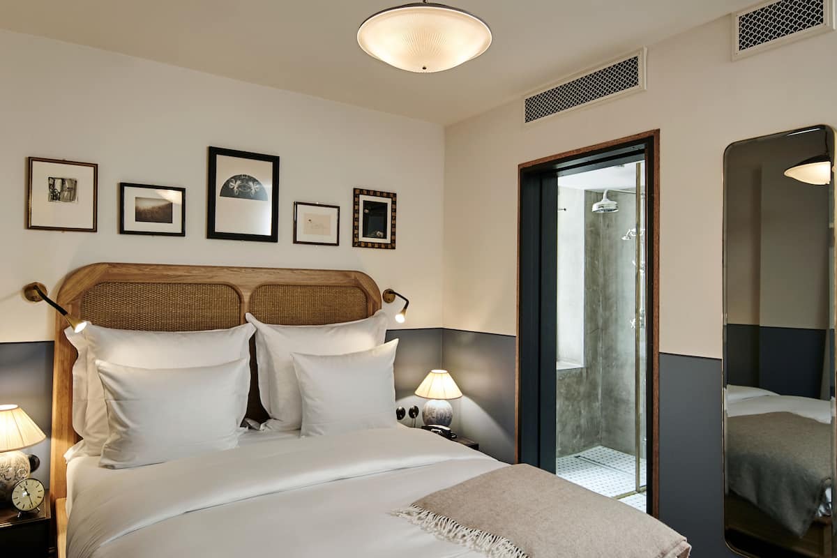 Habitación de hotel con cama, lámparas de noche, cuadros en la pared, un espejo y una puerta que da al baño.