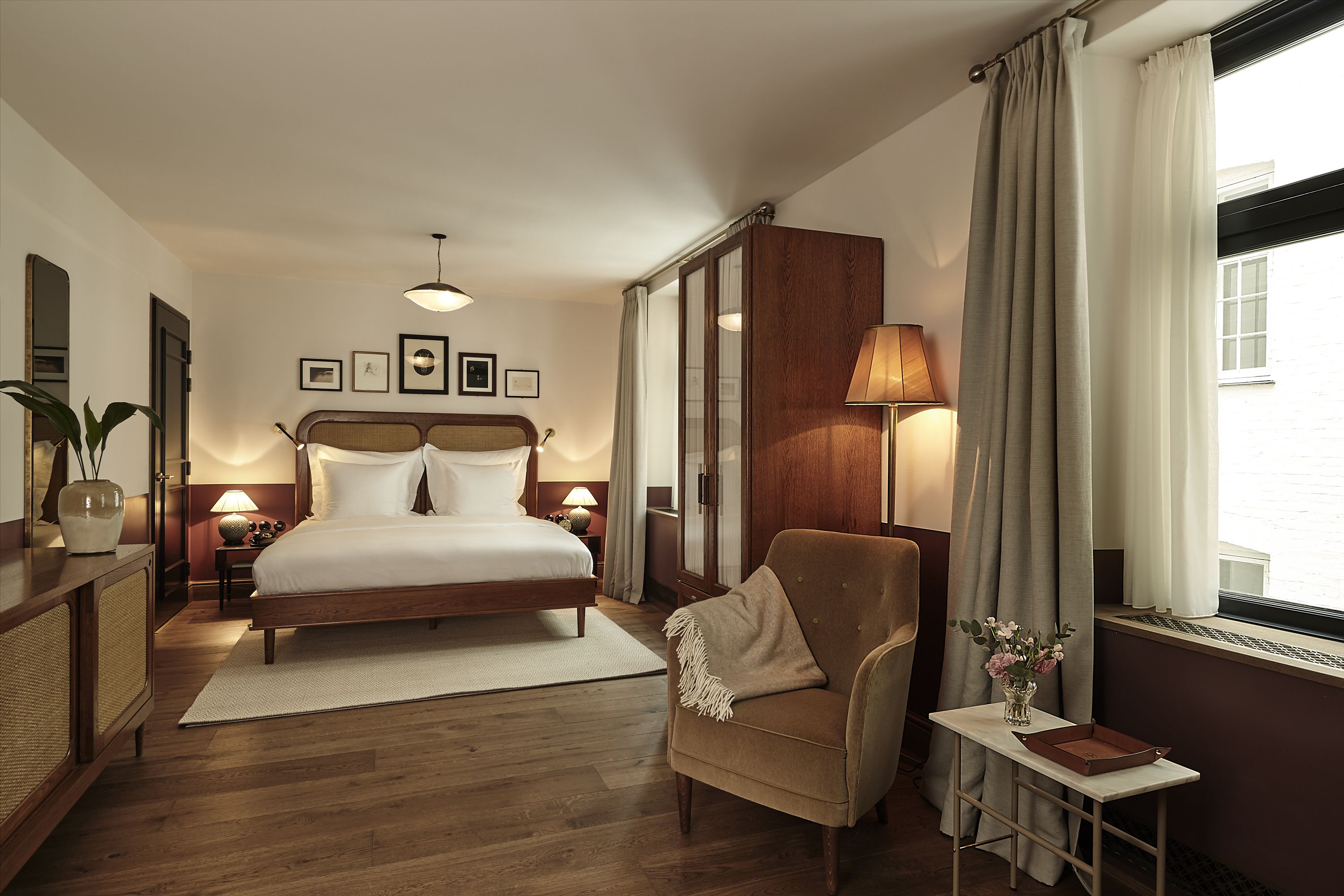 A hotel room with a bed, a chair, a small table with a vase, and a window with curtains.