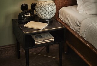 A neatly made bed with white linens, a bedside lamp with a patterned base, a vintage telephone, and a book on a small wooden nightstand.