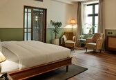 A hotel room with a bed, two armchairs, a side table, a lamp, a potted plant, and a window with curtains.