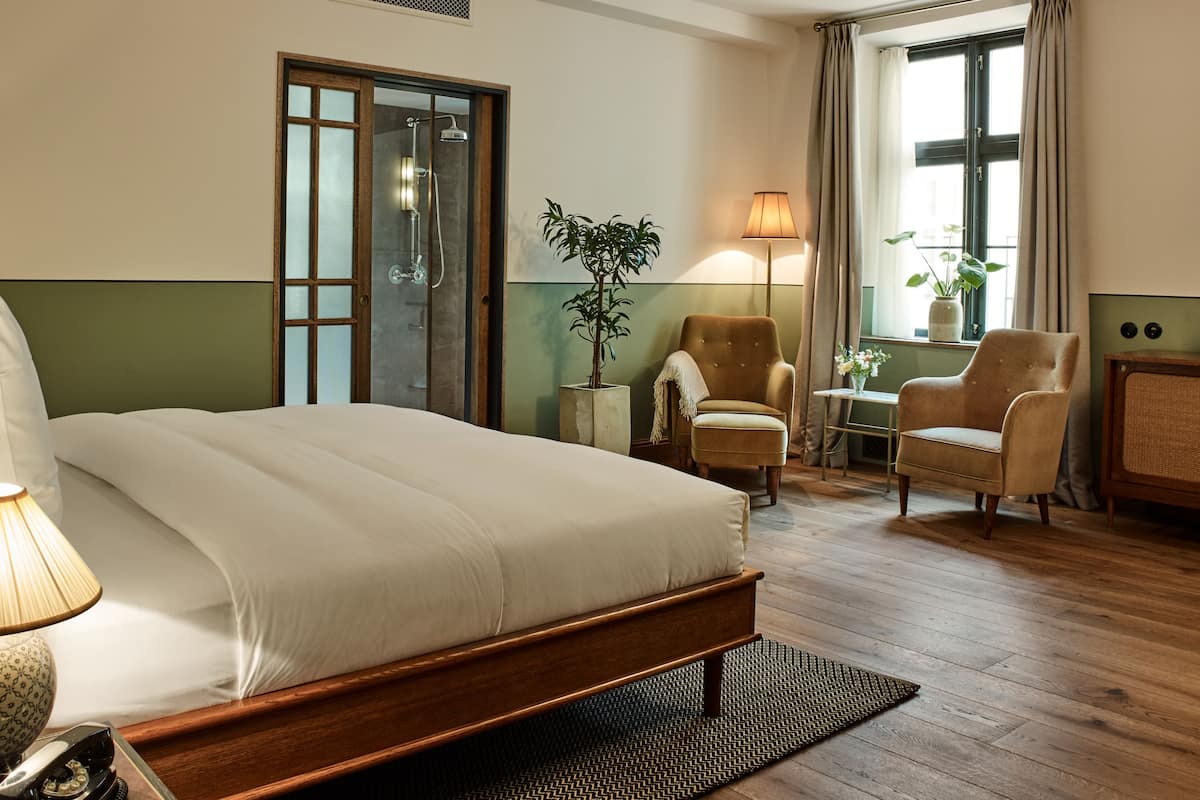 Habitación de hotel con cama, dos sillones, una mesita auxiliar, una lámpara, una planta en maceta y una ventana con cortinas.