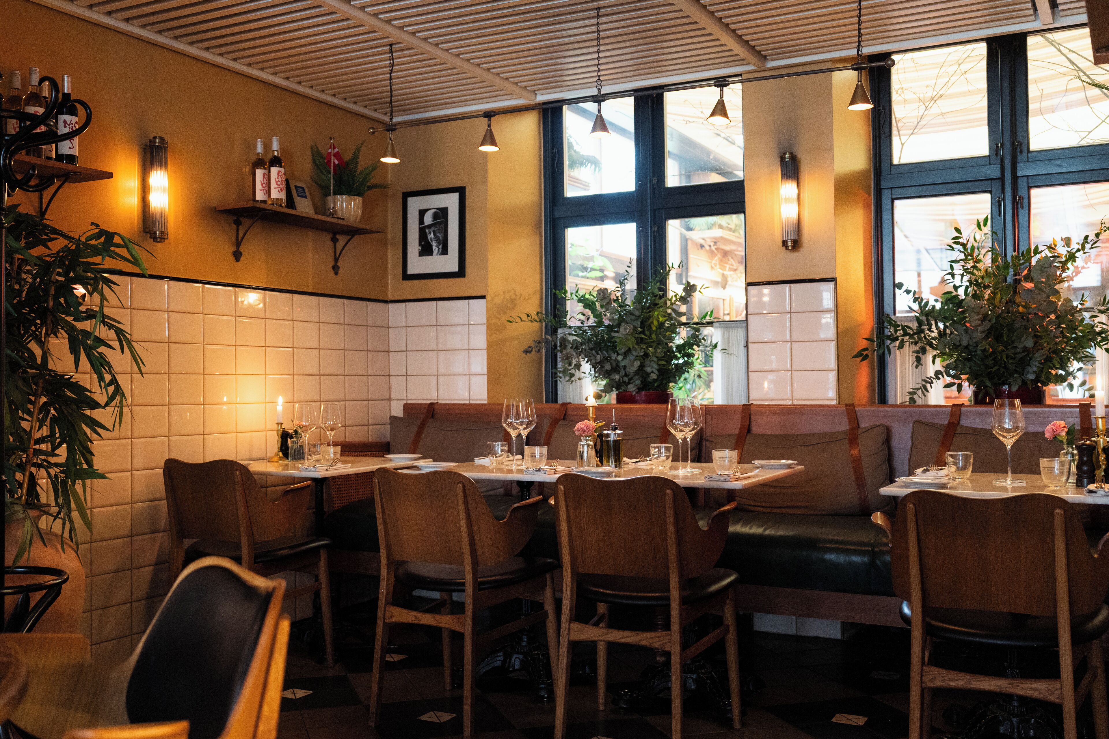 A restaurant interior with wooden booths and tables, potted plants, and wall-mounted shelves with bottles.