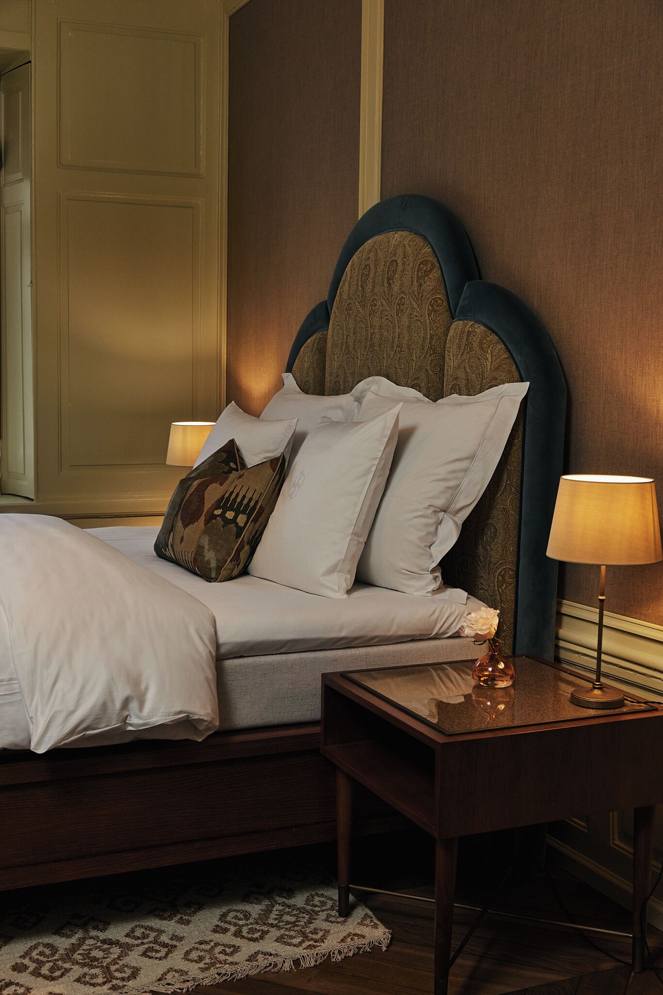 A neatly made bed with white linens and decorative pillows, a bedside table with a lamp, and a patterned rug on the floor.