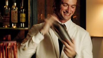 A bartender in a white suit preparing a drink at a bar with shelves of liquor bottles in the background.