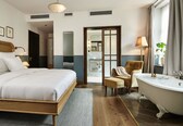 A hotel room with a large bed, a sitting area with a chair and a small table, a bathtub, and a mirror.