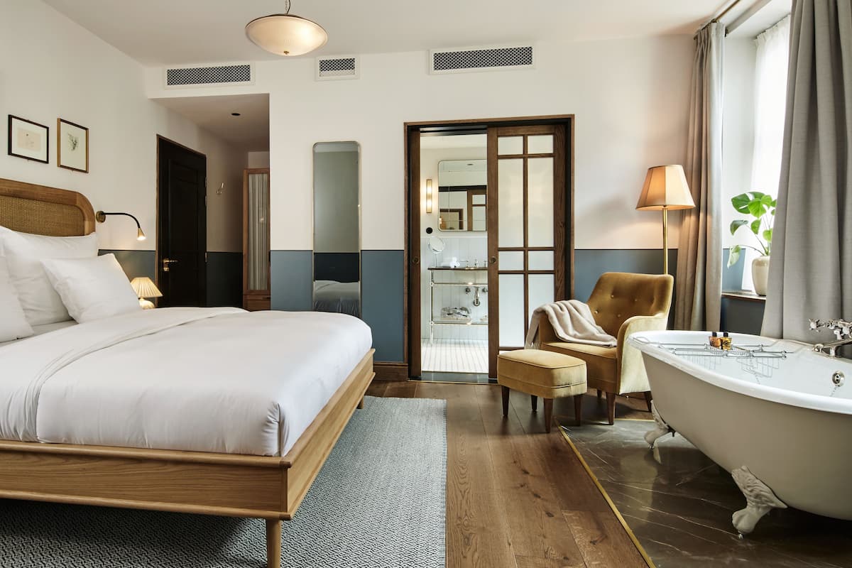 Habitación de hotel con una cama grande, un espacio de descanso con una silla y una mesita, una bañera y un espejo.