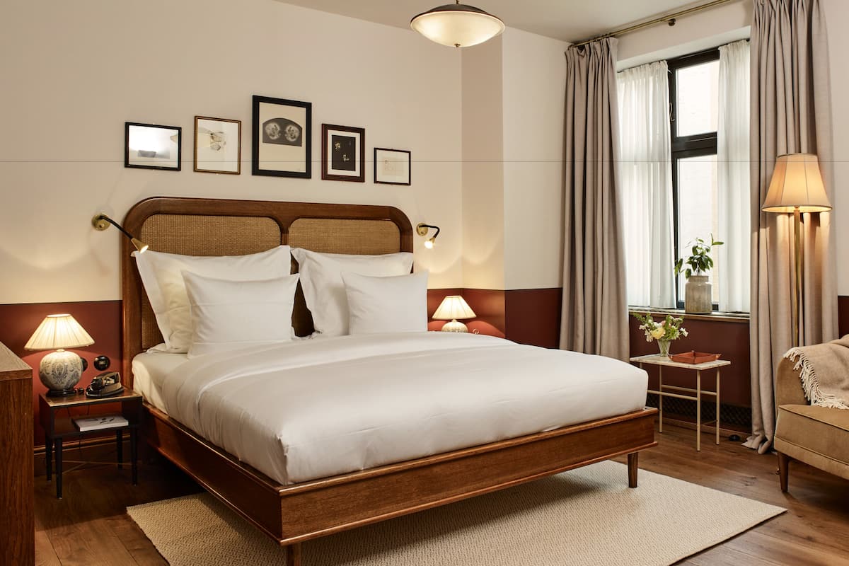 Una cama bien hecha con cabecera de madera, una mesita de noche con lámpara, una silla y una ventana con cortinas.