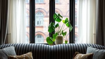 A room with a striped sofa, a potted plant, and a view of a red brick building through a large window with white curtains.
