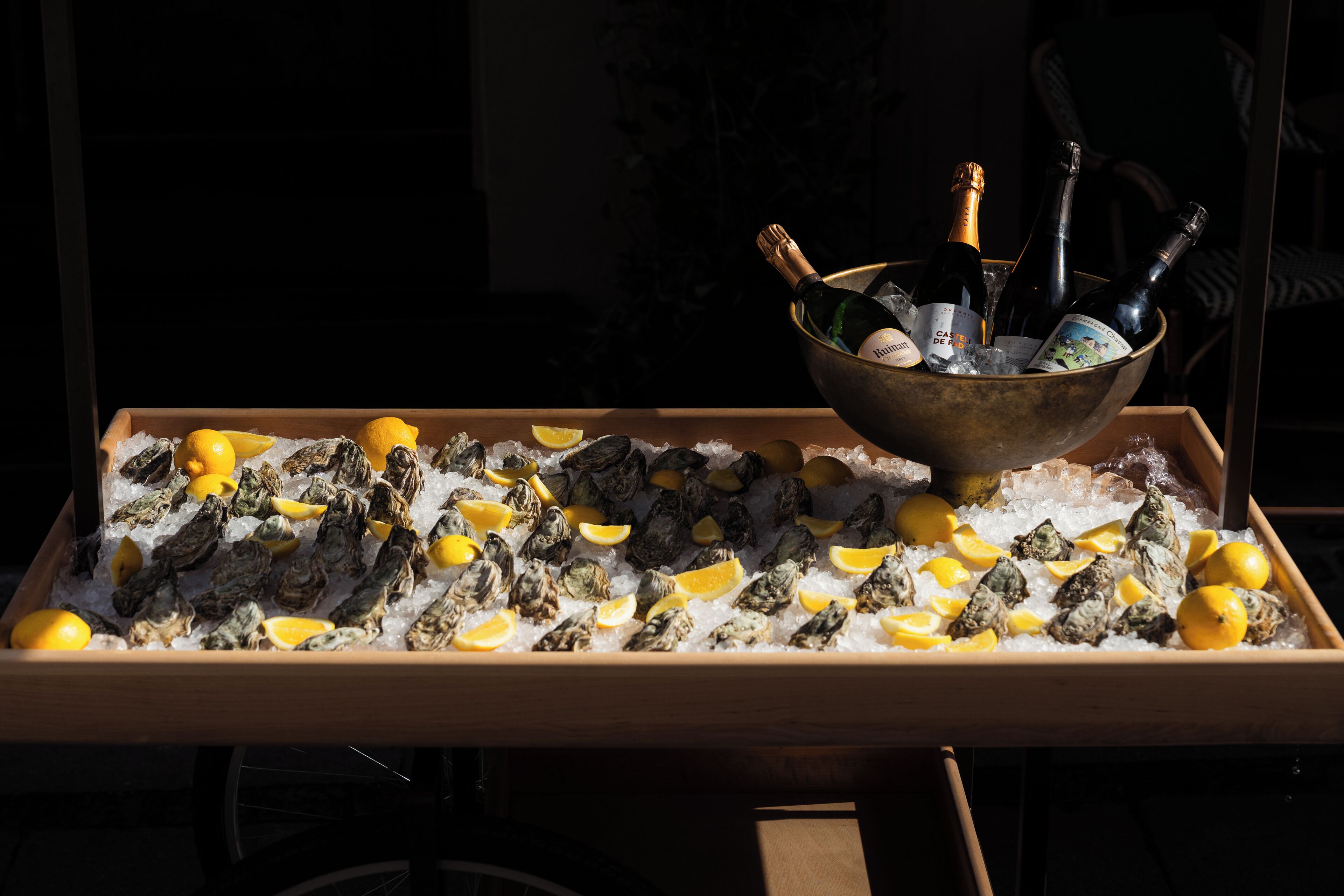 Una bandeja de ostras sobre hielo con rodajas de limón y botellas de champán en un cuenco de metal.