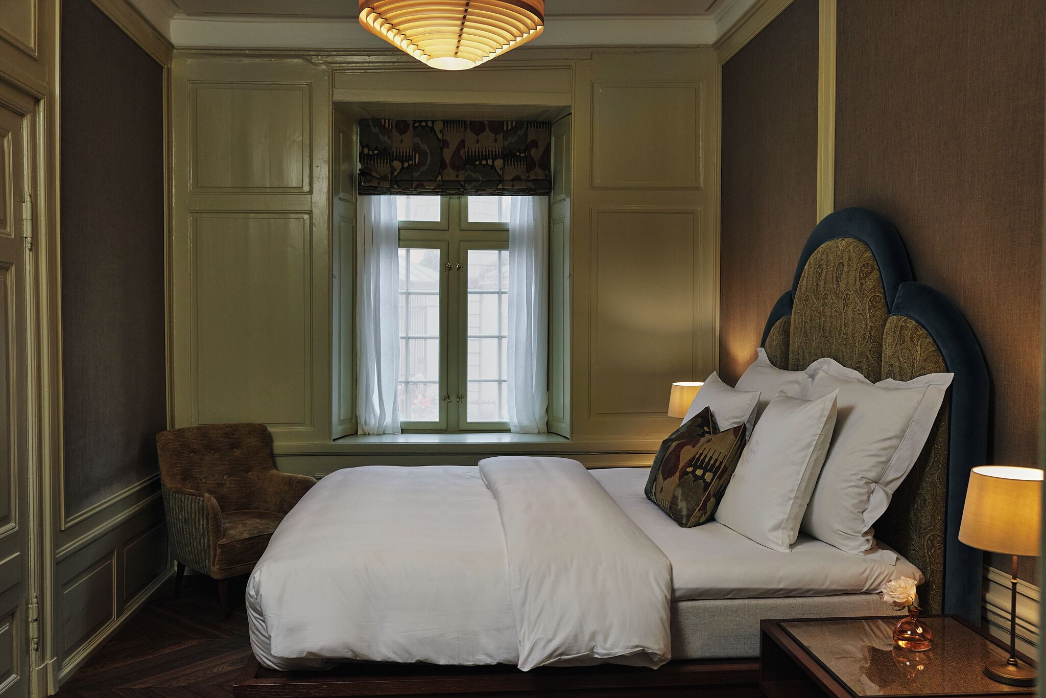A neatly arranged bedroom with a large bed, a chair, a window with a cross, and a lamp on a bedside table.