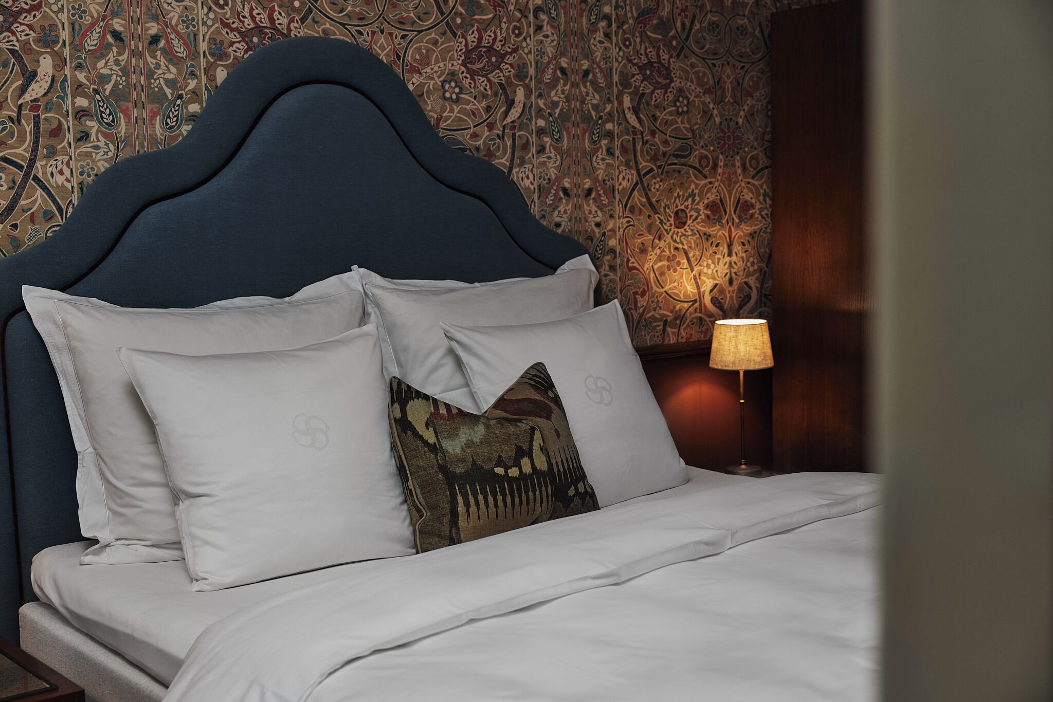 Una cama con cabecera oscura, ropa de cama blanca y almohada estampada.