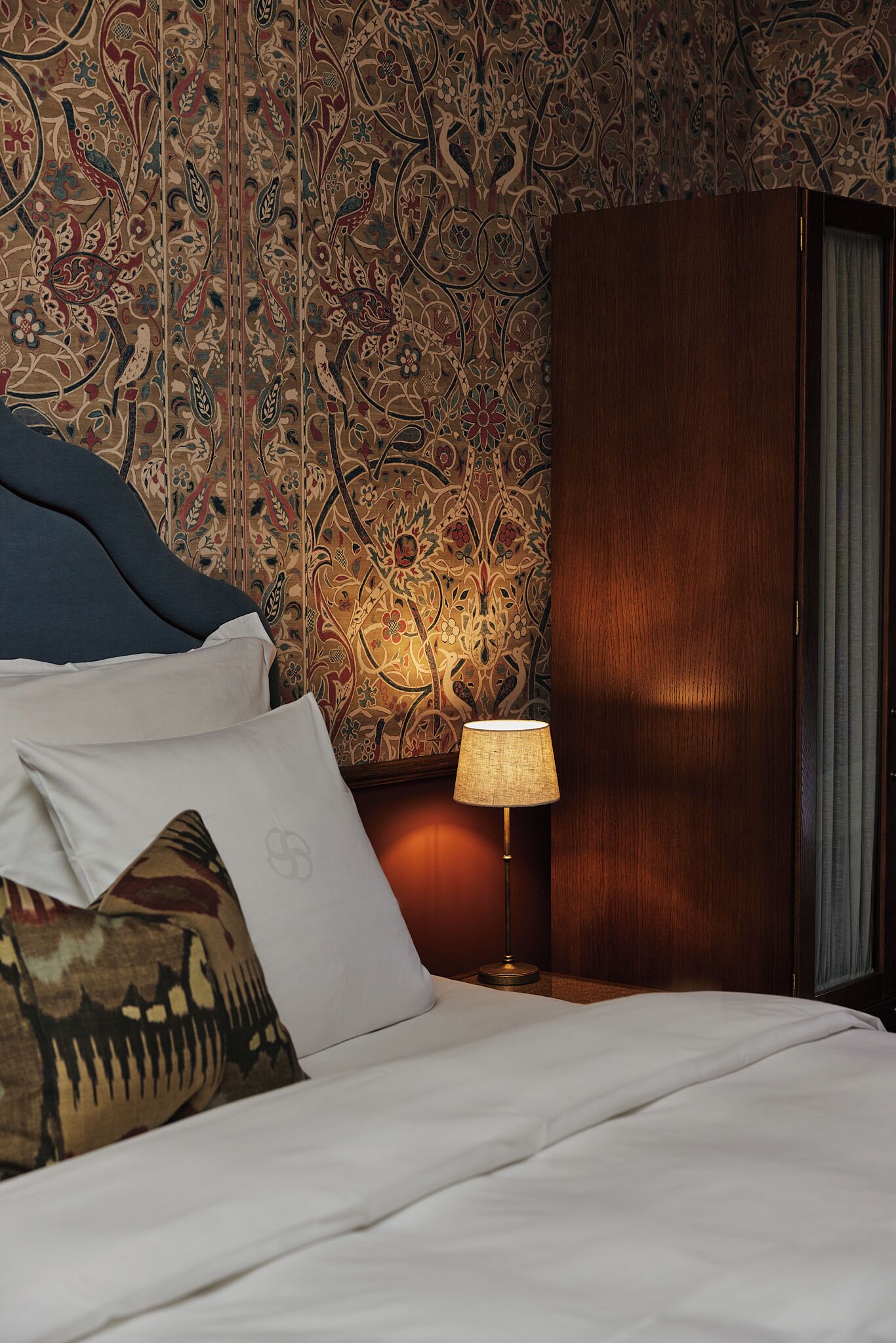 Een net opgemaakt bed met witte lakens en een blauw hoofdbord, een houten garderobe en behang met een patroon.