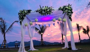 A wedding arch decorated with white drapes and floral arrangements, set against a sunset backdrop. - SeeSea Resort (La-ngu)