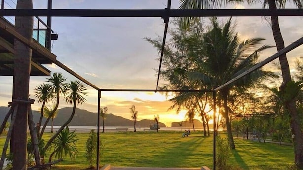 A view from a covered outdoor area with a concrete pathway leading to a grassy field, palm trees, and a sunset over a body of water. - SeeSea Resort (La-ngu)